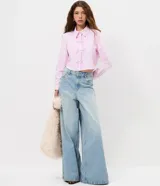 Camisa cropped de manga larga en tejido tricoline, con estampado de rayas verticales finas en color rosa y blanco. Presenta cuello camisero tradicional y cierre frontal decorado con tres lazos de tela. Diseño de corte corto y ajuste recto.
