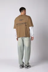 Remera de manga larga con diseño de doble capa, que simula una remera de manga corta color moka sobre una de manga larga blanca. Presenta un pequeño logo gráfico en el pecho y un detalle de etiqueta en el puño.