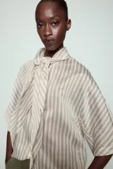 Camisa de corte holgado confeccionada en mezcla de lyocell, con estampado de rayas verticales en tonos neutros y azules. Presenta cuello redondo con lazada frontal, cierre con botones ocultos y mangas amplias por debajo del codo.