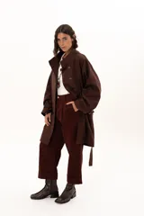 Trench color chocolate con cuello alto, cierre frontal con botones y cinturón ajustable en la cintura. Presenta mangas largas con puños ajustables mediante tiras y hebillas, y bolsillos frontales.