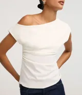 Blusa blanca de corte ajustado, con diseño asimétrico de un solo hombro, manga corta estructurada y detalle drapeado en el lateral de la cintura.