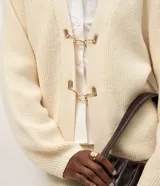 Cardigan de punto color crema con calce oversize, mangas amplias y cierre frontal mediante hebillas metálicas decorativas. Presenta una textura estructurada y puños ajustados.