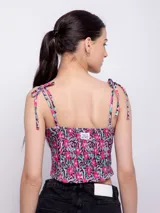 Top corto con escote recto y tirantes finos ajustables mediante nudo en los hombros. Presenta un estampado floral sobre fondo oscuro, con flores rosadas y hojas turquesas.