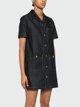 Vestido corto de denim oscuro con cuello camisero, cierre frontal de botones dorados y bolsillos frontales tipo parche con solapa. Presenta costuras en contraste y mangas cortas.