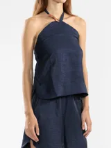 Top halter azul marino confeccionado en lino italiano con hilo metalizado, que aporta un sutil efecto de brillo y una textura natural. El escote halter se ajusta mediante un cordón al cuello, donde se ubica la insignia del tigre Polonio, fácilmente removible al deslizar el cordón.