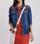 Camisa de jean azul con diseño de arte pintado a mano en color rojo en los hombros y bolsillos frontales con solapa. Cuenta con mangas largas y cierre frontal con botones.