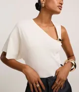 Blusa de punto color crudo, con escote en V y mangas asimétricas: un lado con manga corta y otro musculosa. Tiene un adorno metálico dorado en el bretel.