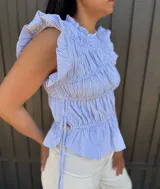 Blusa sin mangas a rayas finas en celeste y blanco, con cuello redondo alto y volados en los hombros. Presenta frunces en el frente, lazos laterales regulables y bajo con ligero vuelo.