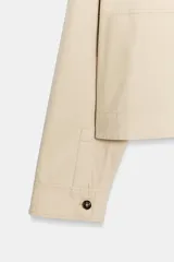 Trench corta de color beige, con cuello solapa, manga larga y cierre frontal con botones oscuros. Presenta bolsillos de vivo en el delantero y un diseño de corte recto.