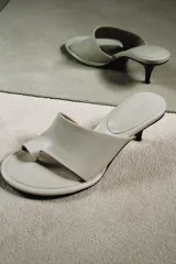 Sandalia tipo mule de tacón alto fino de madera, confeccionada en piel color blanco marfil. Presenta una tira asimétrica con separador para el dedo y punta redonda.