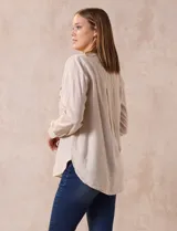 Blusa blanca de lino y rayón, con cuello a la base, escote en V y abertura con botones. Tiene bolsillos en la parte delantera y mangas largas regulables con presilla y botón.