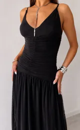 Vestido largo negro con escote profundo en V, tirantes finos y diseño ajustado en la cintura con detalle fruncido, que se abre en una falda fluida y amplia.