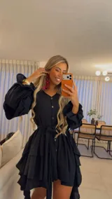 Vestido camisero corto de muselina, color negro, con mangas largas abullonadas, falda con volados en cascada y cierre frontal con botones de coco. Incluye lazo ajustable en la cintura.