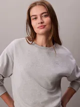 Buzo de felpa francesa gris claro, de cuello redondo y manga larga, con puños y dobladillo acanalados. Presenta un diseño holgado y lleva el logo retro de Calvin Klein bordado discretamente en el pecho.