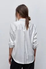 Camisa blanca de calce relajado con acabado satinado y brillo sutil, cuello clásico, cierre frontal con botones y mangas largas con puños.