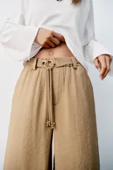 Pantalón recto color beige de tiro medio, con cintura elástica, bolsillos delanteros y cinturón ajustable con hebilla metálica dorada.
