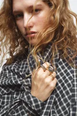 Pack de tres anillos dorados. El primero es un anillo sello con piedra central marrón rodeada de pequeños brillos oscuros. El segundo es un anillo sello liso con borde inferior de brillos oscuros. El tercero es una banda fina con una hilera de pequeñas esferas doradas en la parte inferior.