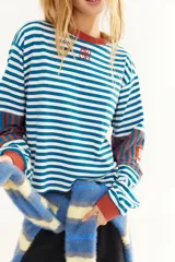 Remera de manga larga con corte boxy, confeccionada en algodón. Presenta un diseño de rayas horizontales en azul y blanco, con detalles de rayas verticales en tonos rojizos en los codos y puños en contraste. Incluye logo bordado en el pecho y tajos laterales.