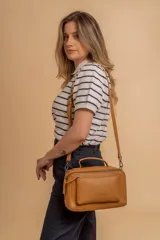 Bandolera de diseño rectangular en color suela, confeccionada en material sintético. Cuenta con un asa superior fija, correa larga ajustable y extraíble, y dos bolsillos frontales con cierre metálico, uno de ellos con diseño superpuesto. Presenta el logo de la marca grabado en el frente.