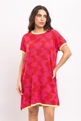 Vestido corto tejido de punto con diseño geométrico abstracto en tonos rojo y fucsia, cuello redondo, mangas cortas y bolsillos laterales, con detalles en amarillo en el cuello, puños y ruedo.