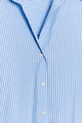 Camisa celeste con rayas verticales blancas, de cuello solapa y escote pico. Manga larga acabada en puño, bajo asimétrico y cierre frontal con botones.