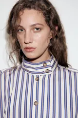 Camisa de manga larga con cuello subido y trabillas en los hombros. Presenta un diseño de rayas verticales en tonos azul, beige y marrón, con cierre frontal de botones.