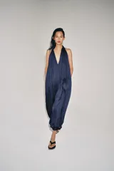 Mono largo satinado de color azul marino, con cuello halter, escote en V profundo y espalda descubierta. Presenta diseño plisado y bajo abullonado con terminación en puño.
