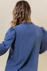 Camisa azul de calce relajado con bordado floral blanco en el frente. Presenta mangas largas amplias con ligero fruncido en el hombro y cierre frontal con botones.