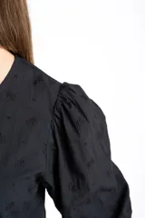 Blusa negra de algodón con diseño de broderie, mangas tres cuartos abullonadas, cierre frontal con botones y detalle de cordón ajustable en la cintura.