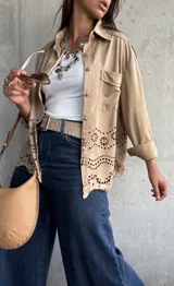 Sobrecamisa de gamuza color beige, con cuello camisero, cierre frontal con botones metálicos y bolsillo en el pecho. Presenta un diseño con calados florales y bordes ondulados en la parte inferior.