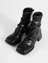 Botas de caña media en color negro, confeccionadas en cuero sintético con acabado ligeramente arrugado. Presentan una puntera cuadrada, cierre posterior y un taco cuadrado de altura media con textura estriada.
