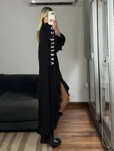 Vestido largo color marrón, con escote en V, mangas largas con puños y detalle de encaje en el pecho. Presenta un diseño asimétrico con volados en el ruedo y una abertura frontal.