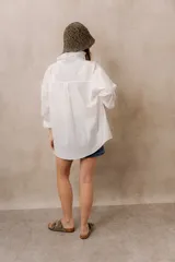 Camisa blanca de calce amplio, confeccionada en tejido liviano con sutiles rayas verticales, cuello clásico, mangas largas con hombros caídos y abotonadura frontal.