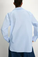 Camisa de popelín confeccionada en algodón, con cuello solapa, escote en pico y cierre frontal con botones. Presenta mangas largas con puños y un bajo redondeado asimétrico.