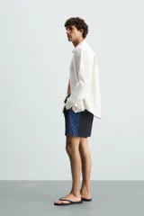 Bañador largo negro con cintura elástica ajustable mediante cordón. Presenta un panel lateral con estampado de cuadros en tonos azul y negro. Incluye bolsillos frontales y un bolsillo trasero con solapa.