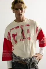 Camiseta boxy fit de cuello redondo y manga por debajo del codo. Presenta un diseño bicolor en tonos blanco off-white y rojo, con estampado y parches frontales con efecto lavado vintage y franjas en las mangas.
