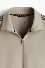 Campera deportiva de color gris topo, con cuello alto, cierre frontal bidireccional y bolsillos laterales. Presenta franjas laterales negras en las mangas y puños y bajo de punto acanalado.