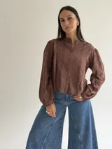 Blusa de brodery color marrón, con cuello mao, cierre frontal con botones forrados y mangas largas abullonadas con puños elásticos. Presenta detalles de puntillas verticales en el frente.