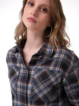 Camisa de corte cropped y diseño holgado, confeccionada en tela de franela con estampado a cuadros en tonos negro, azul y naranja. Presenta cuello camisero, cierre frontal con botones metálicos, mangas largas con puños abotonados y bolsillos frontales en el pecho.