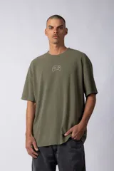 Remera de corte oversize color verde oliva, con cuello redondo y mangas cortas con hombros caídos. Presenta un bordado minimalista de un joystick en el centro del pecho y una etiqueta aplicada en la parte superior de la espalda.