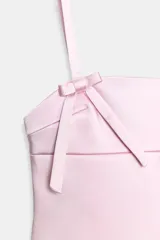 Vestido corto de satén en color rosado, con escote recto y tirantes finos. Presenta un diseño con detalle de lazos decorativos en el delantero y cierre de cremallera oculto en la espalda.