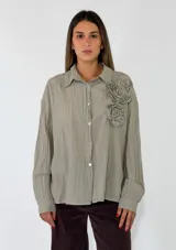 Camisa de corte oversize color beige, con cuello clásico, cierre frontal de botones y detalle de flores de tela aplicadas en el pecho.