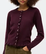 Cardigan de punto color bordó con cuello redondo, cierre frontal de botones de nácar y detalle de volados en la tapeta de botones y el ruedo. Presenta mangas largas con puños ajustados.