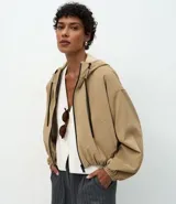 Campera cortavientos de color beige con diseño balonê, capucha con cordones ajustables, cierre frontal de cremallera y puños con elástico. Presenta un corte holgado y moderno.