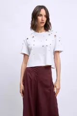 Remera blanca de algodón con cuello redondo y mangas cortas. Presenta apliques metálicos y aberturas circulares en los hombros y el frente.