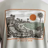 Remera verde con estampa en el pecho de una tienda de surf, palmeras y un atardecer.