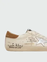 Zapatillas Golden Goose modelo Super-Star de cuero napa color blanco, con la icónica estrella lateral laminada en plateado, talón de gamuza marrón y efecto desgastado artesanal. Presenta inscripciones y detalles escritos a mano en los laterales y la lengüeta.