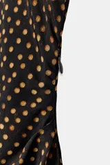 Vestido midi de corte asimétrico con escote barco, una manga corta y un hombro descubierto. Presenta un diseño drapeado en el lateral y estampado de lunares dorados sobre fondo negro.