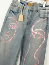 Jean de corte wide leg en denim azul claro, intervenido a mano con ilustraciones abstractas de rostros en líneas color rosa y blanco. Presenta tiro medio-alto y diseño de cinco bolsillos.