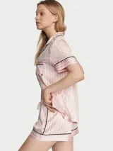 Pijama corta de satín con diseño de rayas verticales rosadas y blancas, ribetes negros en cuello, solapa, bolsillo y dobladillos. Consta de una camisa de manga corta con botones frontales y bolsillo en el pecho, y un short a juego con cintura elástica.